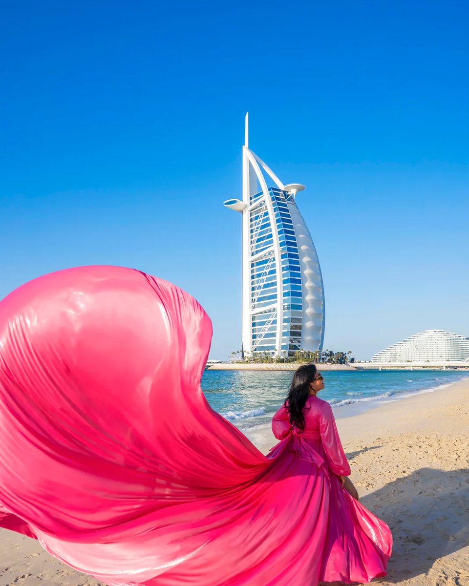 Woman in a hot-pink flying dress on the sand facing the Burj Al Arab across the sea