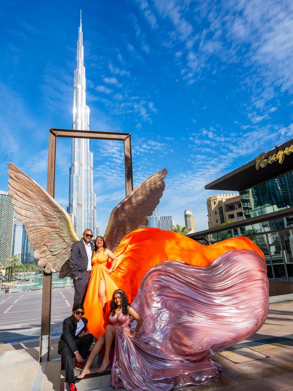 Family of four at the bronze wings sculpture, orange and metallic pink gowns fanned out below the Burj Khalifa