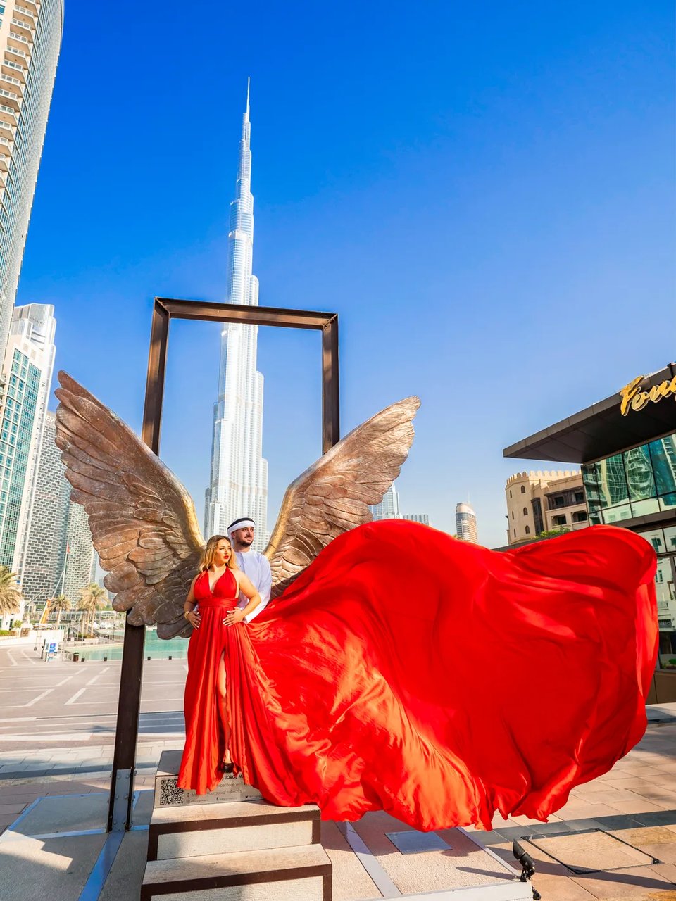 Couple at the bronze wings sculpture as her red flying gown sweeps sideways, Burj Khalifa above