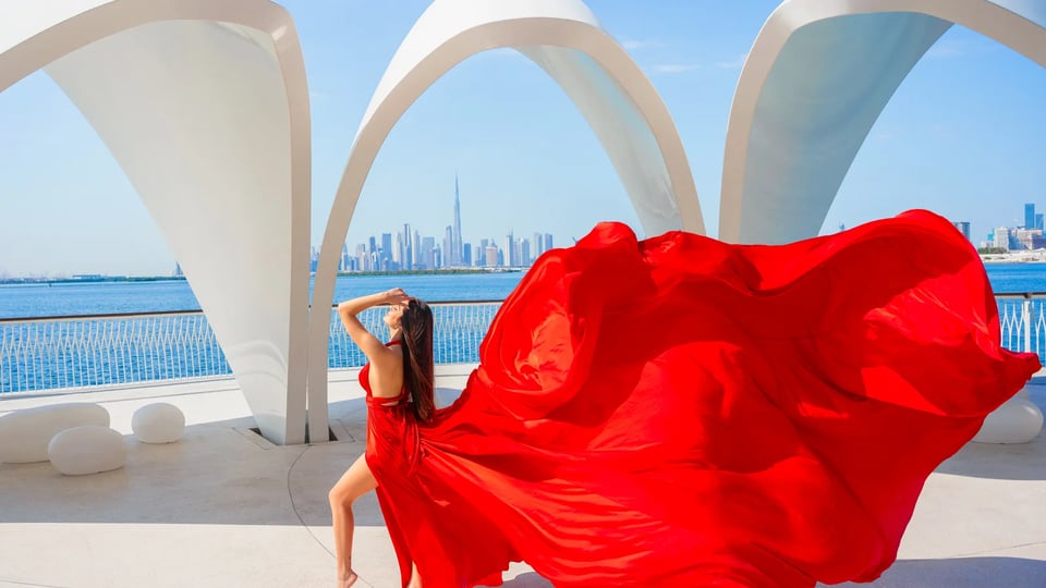 Woman in a red flying dress posing beneath the white arches at Dubai Creek Harbour with the city skyline across the water