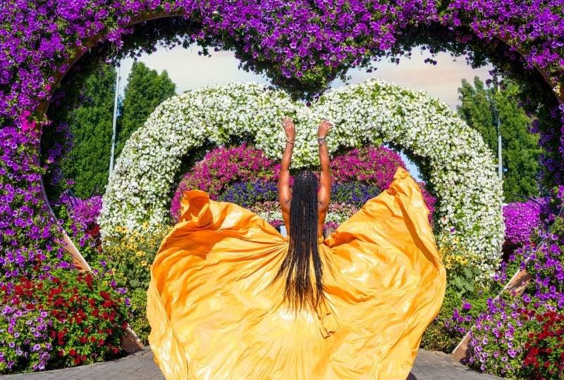 Woman in a golden flying dress raising her arms beneath the heart-shaped flower arches at Dubai Miracle Garden